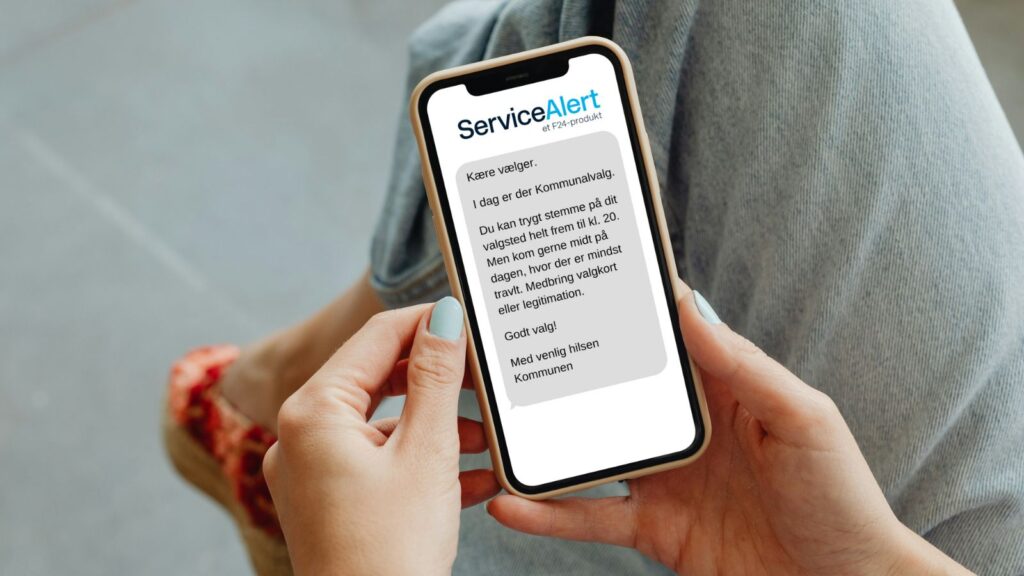 En person holder en smartphone, der viser en dansk besked fra ServiceAlert om kommunalvalg, som opfordrer modtageren til at stemme inden kl. 20. Personen har jeans og røde sko på.