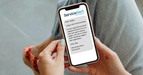 En person holder en smartphone, der viser en dansk besked fra ServiceAlert om kommunalvalg, som opfordrer modtageren til at stemme inden kl. 20. Personen har jeans og røde sko på.