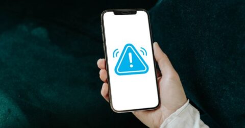 En hånd holder en smartphone med et blåt advarselssymbol på skærmen mod en mørkegrøn baggrund.