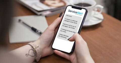 Person håller mobil med ServiceAlert-meddelande om driftstörning via SMS, med anteckningsbok och kaffe i bakgrunden.