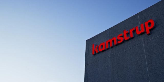 Kamstrups byggnad med företagets logotyp på fasaden mot klarblå himmel.